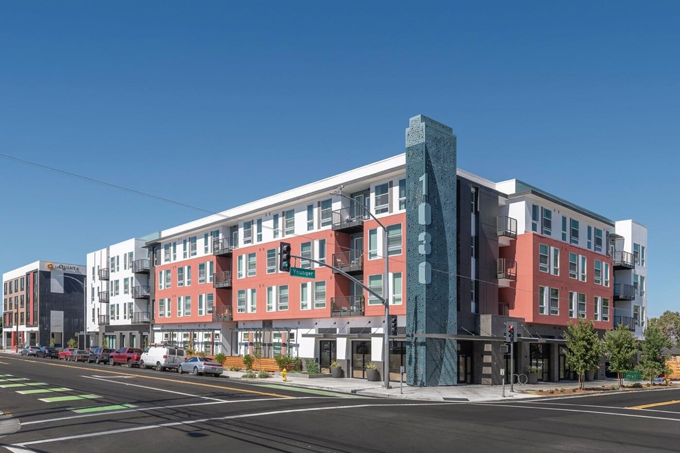 A modern four-story apartment building with red and white exterior, large windows, balconies, and a tall blue-gray vertical sign displaying "1030" at the corner. Cars line the street along a sunny, clear sky.