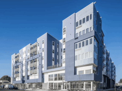 Modern multi-story apartment building with large windows, balconies, and a blue and white facade under a clear blue sky. Ground floor features glass storefronts.