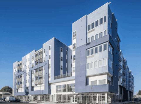 Modern multi-story apartment building with large windows, balconies, and a blue and white facade under a clear blue sky. Ground floor features glass storefronts.