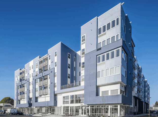 Modern multi-story apartment building with large windows, balconies, and a blue and white facade under a clear blue sky. Ground floor features glass storefronts.